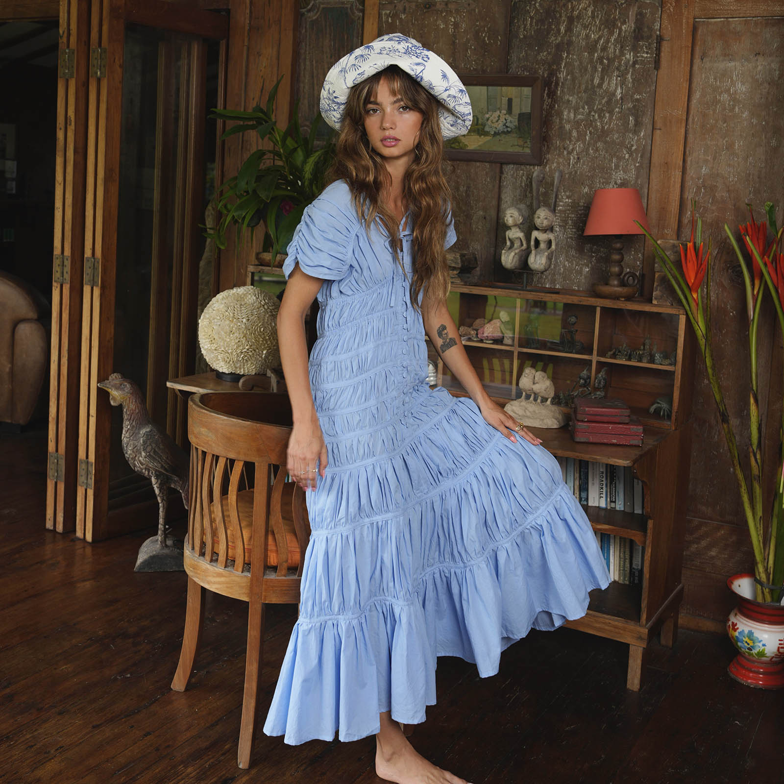 Woman in a blue dress and hat standing in a room with wooden furniture and decor.