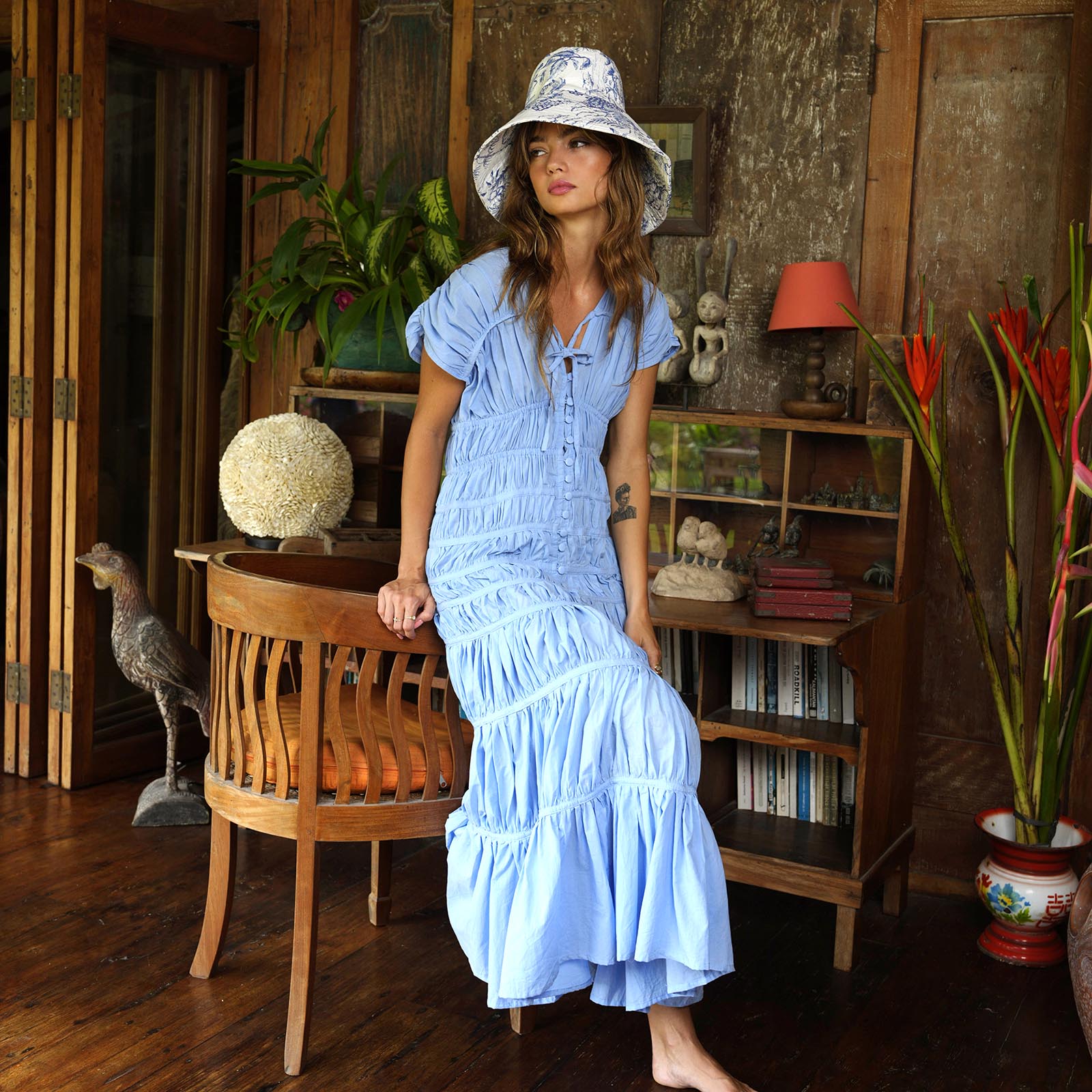 Woman in a blue dress and sun hat standing in a room with wooden furniture and plants.