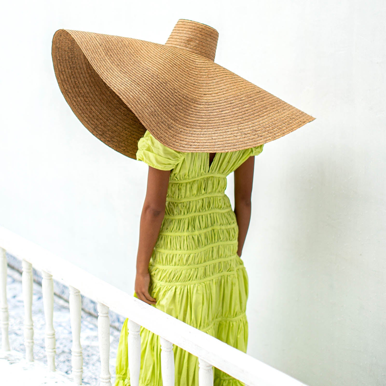 Person wearing a large straw hat and green dress by a white railing.