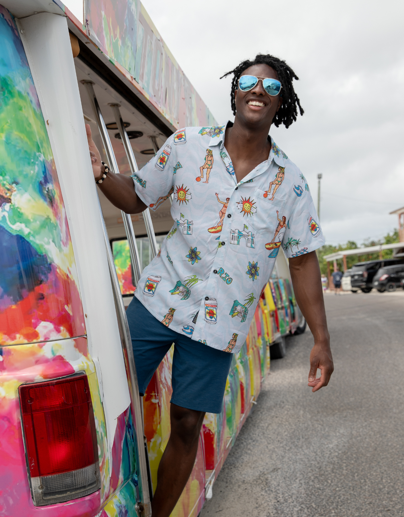 Man in a colorful shirt and sunglasses standing next to a vibrant, artfully painted vehicle.