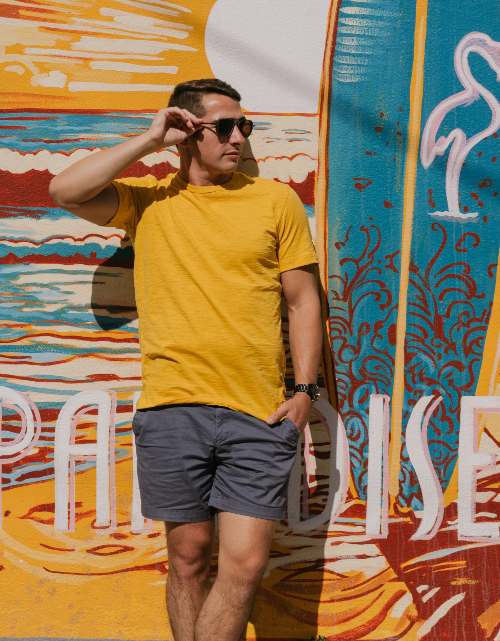 Man in yellow shirt and sunglasses holding a surfboard in front of a colorful mural.