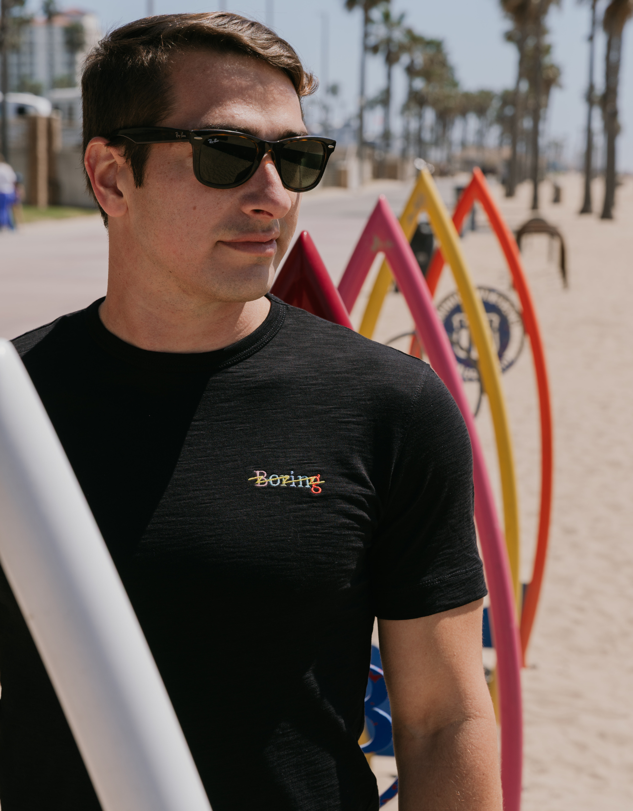 Man wearing sunglasses and a black t-shirt with a logo, standing on a beach with surfboards in the background.
