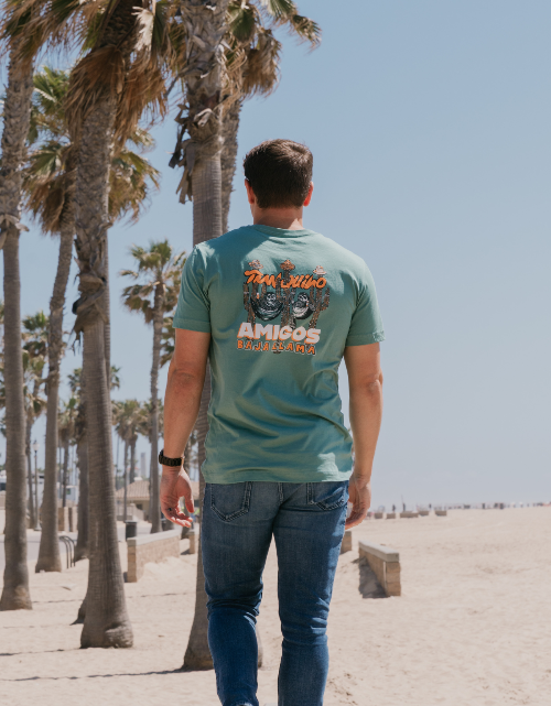 Man walking on a beach with palm trees, wearing a green t-shirt with text and graphics.
