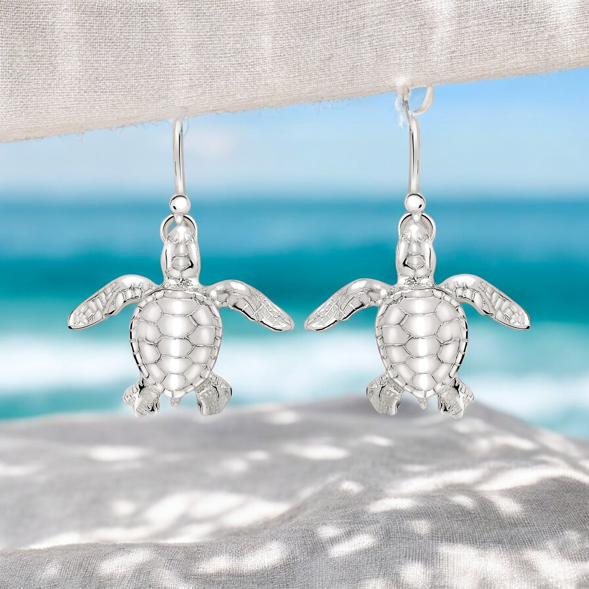 Silver turtle earrings on a textured surface with a blurred beach background