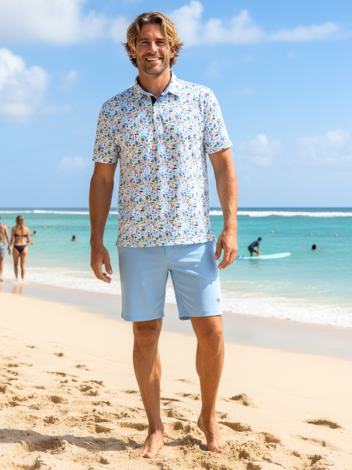 Man standing on a beach wearing a patterned shirt and light blue shorts with a clear blue sky and ocean in the background.