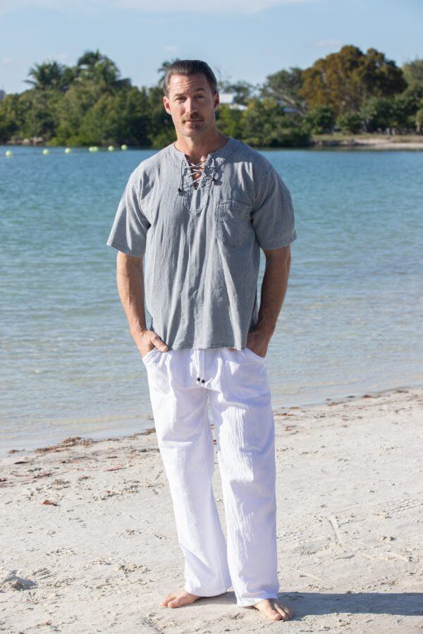 Man standing on a beach wearing a gray shirt and white pants with trees and water in the background.