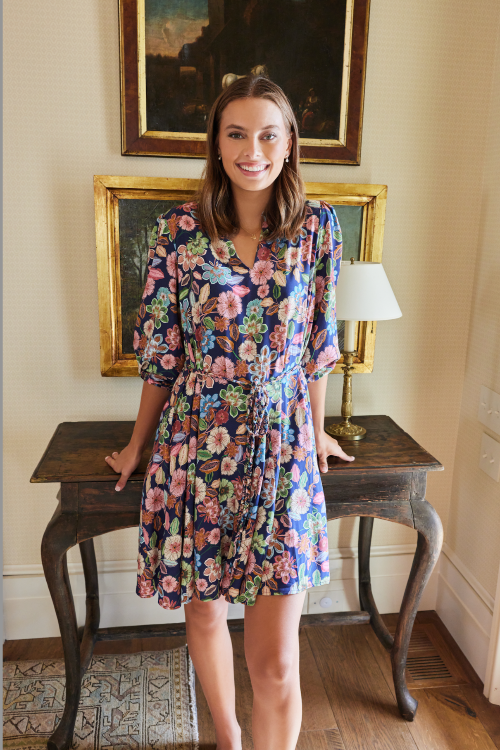 Woman in a floral dress standing in a room with wooden table and framed artwork.