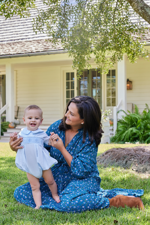 Woman in a blue floral dress holding a child in a white outfit outdoors.