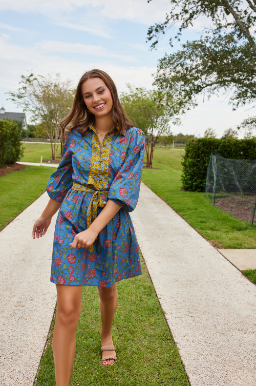 Woman in a blue floral dress standing on a sidewalk with greenery in the background