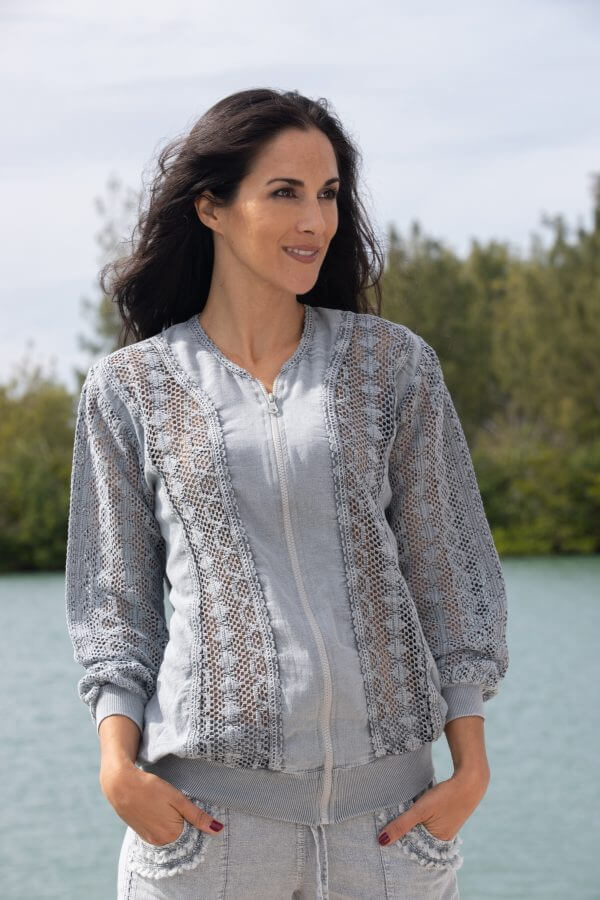 Woman wearing a light gray lace cardigan over a white shirt, standing by a body of water with greenery in the background.