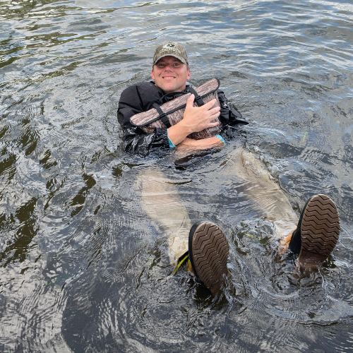 Person in water holding a large bag, wearing a cap and boots.