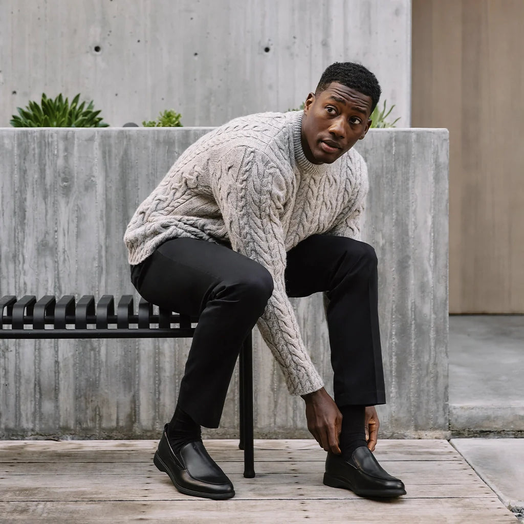 Man sitting on a bench wearing a cable knit sweater and black shoes, with a concrete wall and plants in the background.