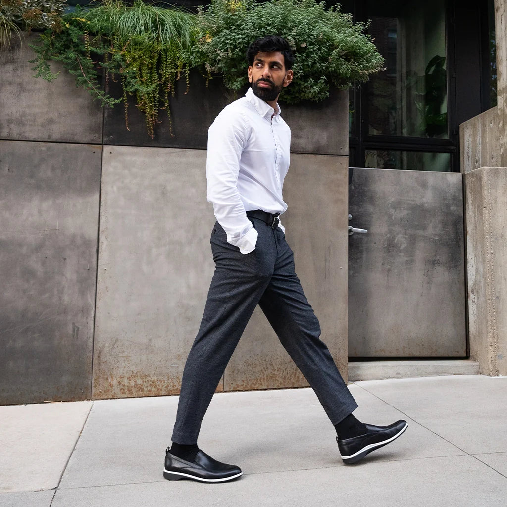 Man in a white shirt and gray pants walking outdoors near a building with plants.