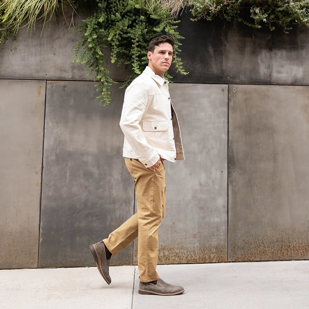 Man in a white jacket and beige pants standing against a metallic wall with greenery.