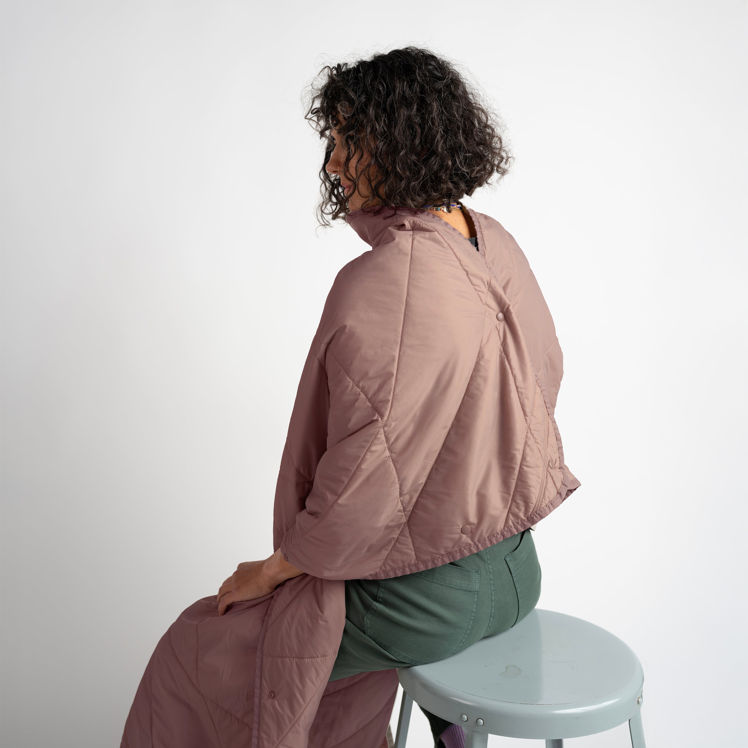 Person wearing a pink quilted jacket sitting on a white stool against a white background