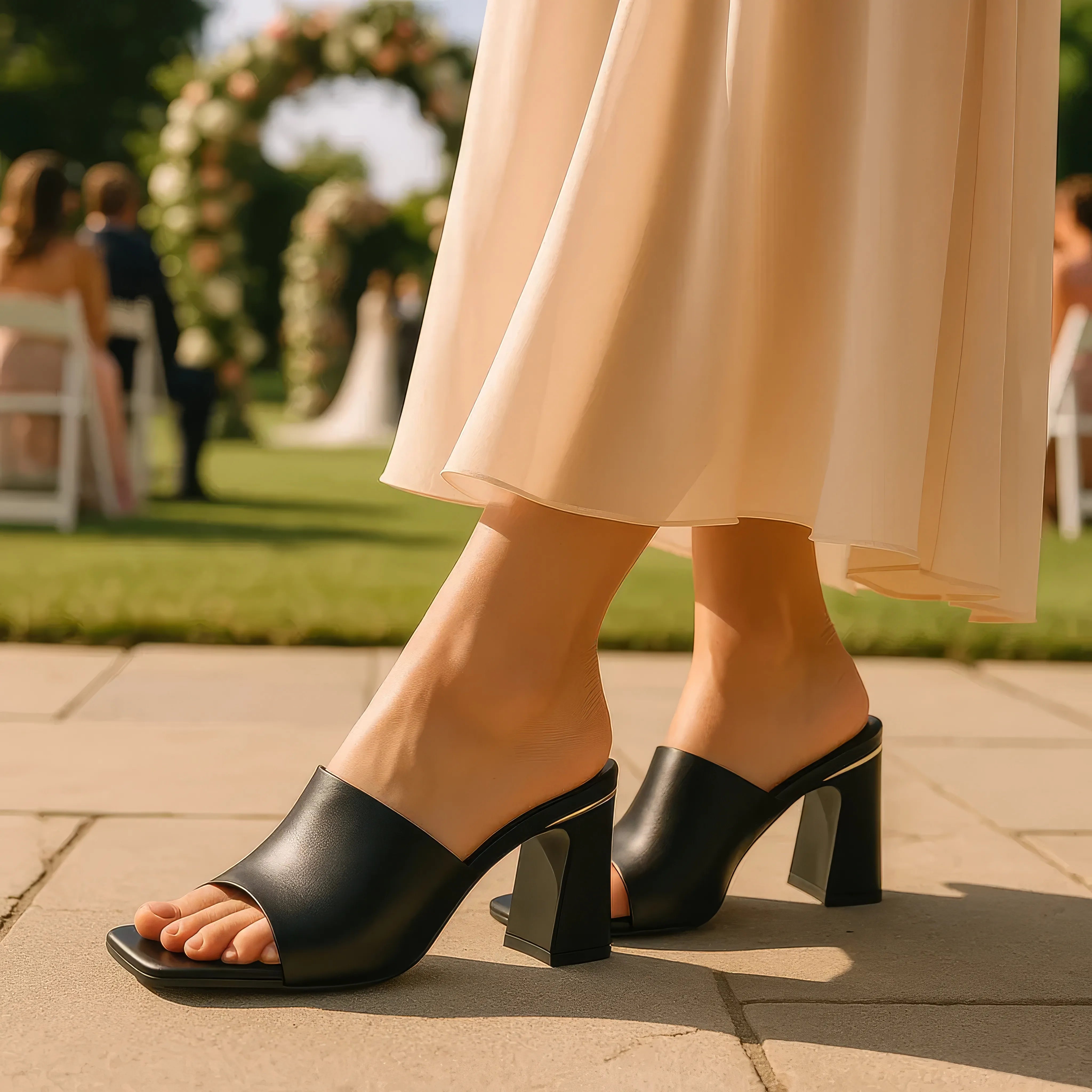 Black open-toe high-heeled shoes worn outdoors with a blurred wedding scene in the background.