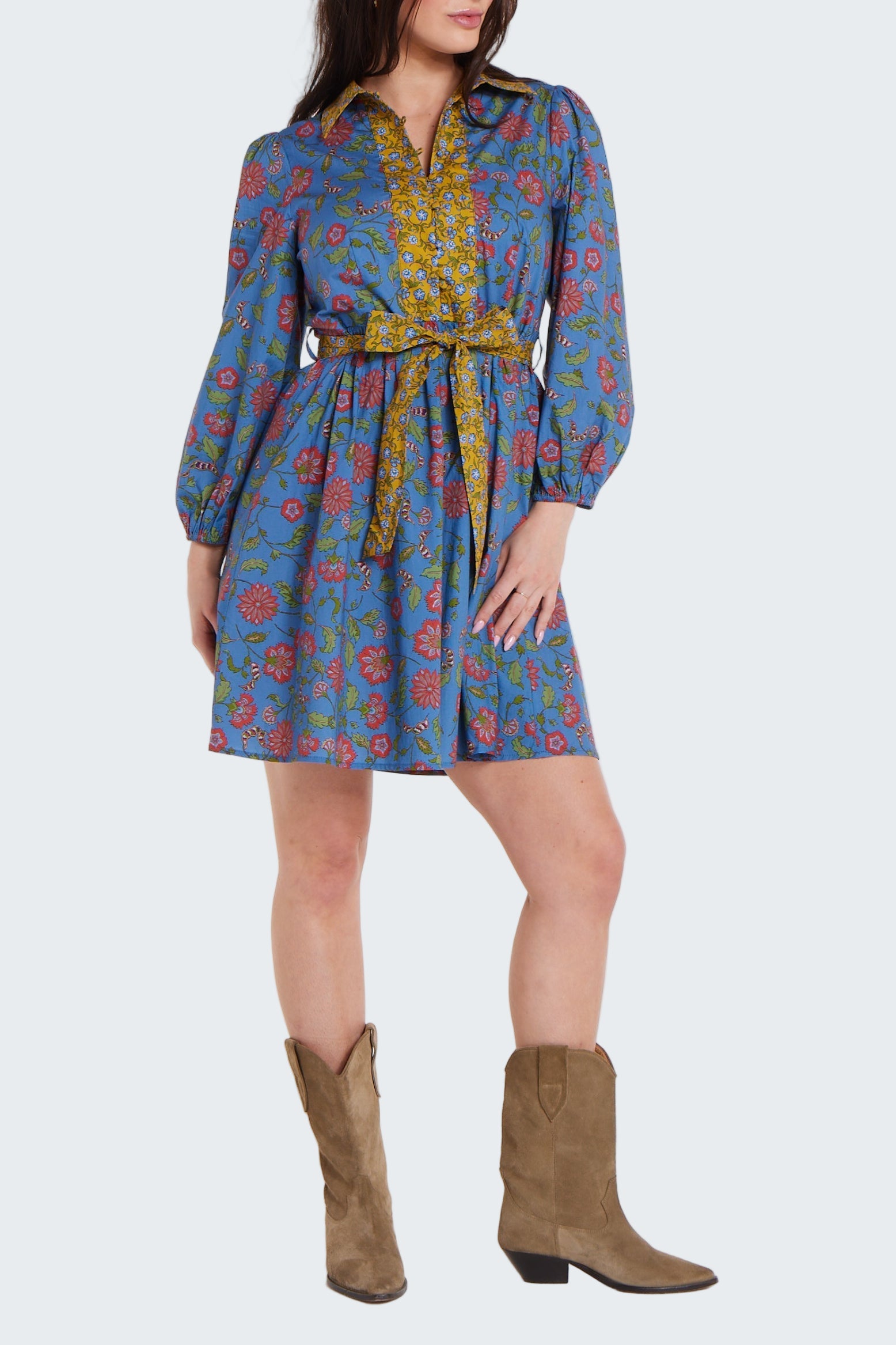 Woman wearing a patterned dress with long sleeves and a belt, paired with brown boots on a white background.