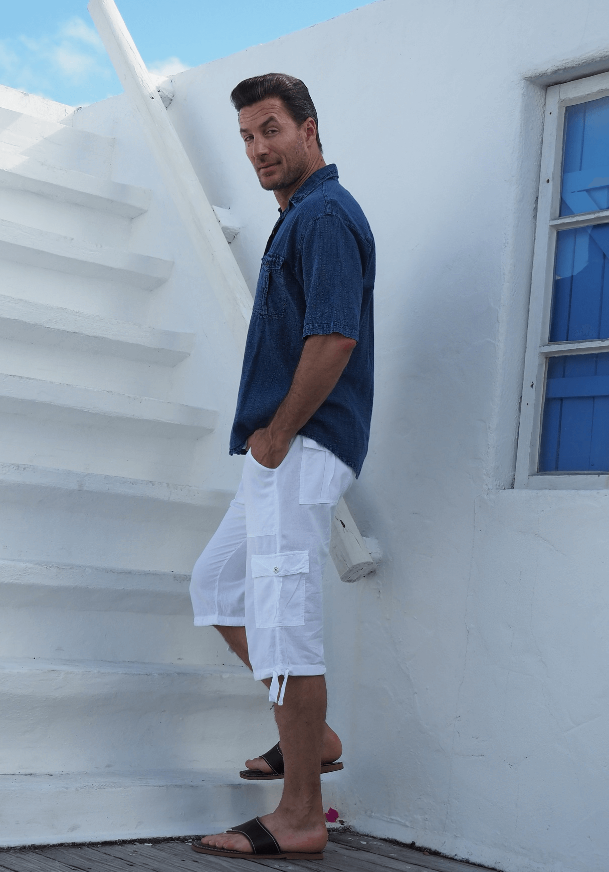 Man in a blue shirt and white shorts standing on a white staircase with a blue door in the background.