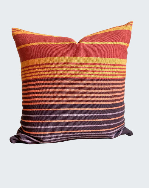 Striped pillow with red, orange, and purple colors on a wooden stool against a white wall.