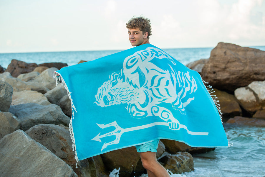 Person holding a turquoise towel with a white graphic design by the ocean.