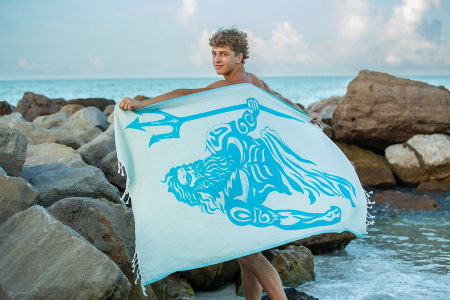 Person holding a towel with a design of a trident and figure on a rocky beach.