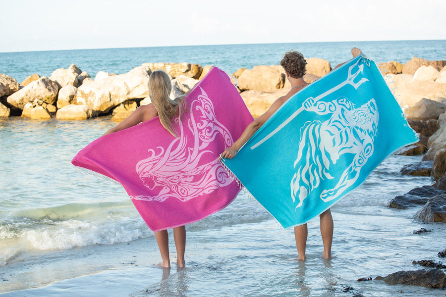 Two people holding pink and blue towels with animal designs on a rocky beach.