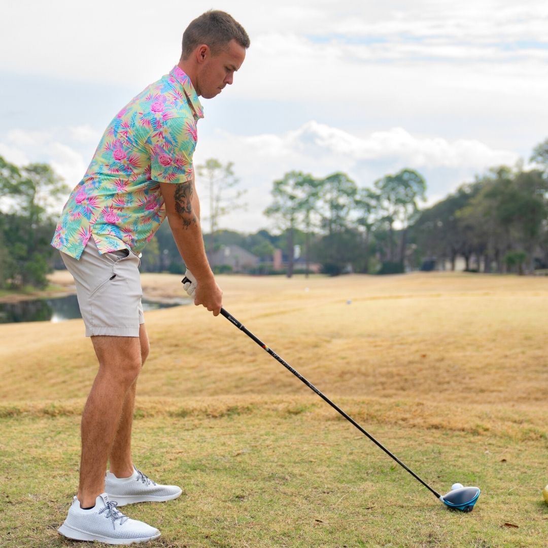 Man in a colorful shirt and white shorts playing golf on a grassy field with trees in the background.