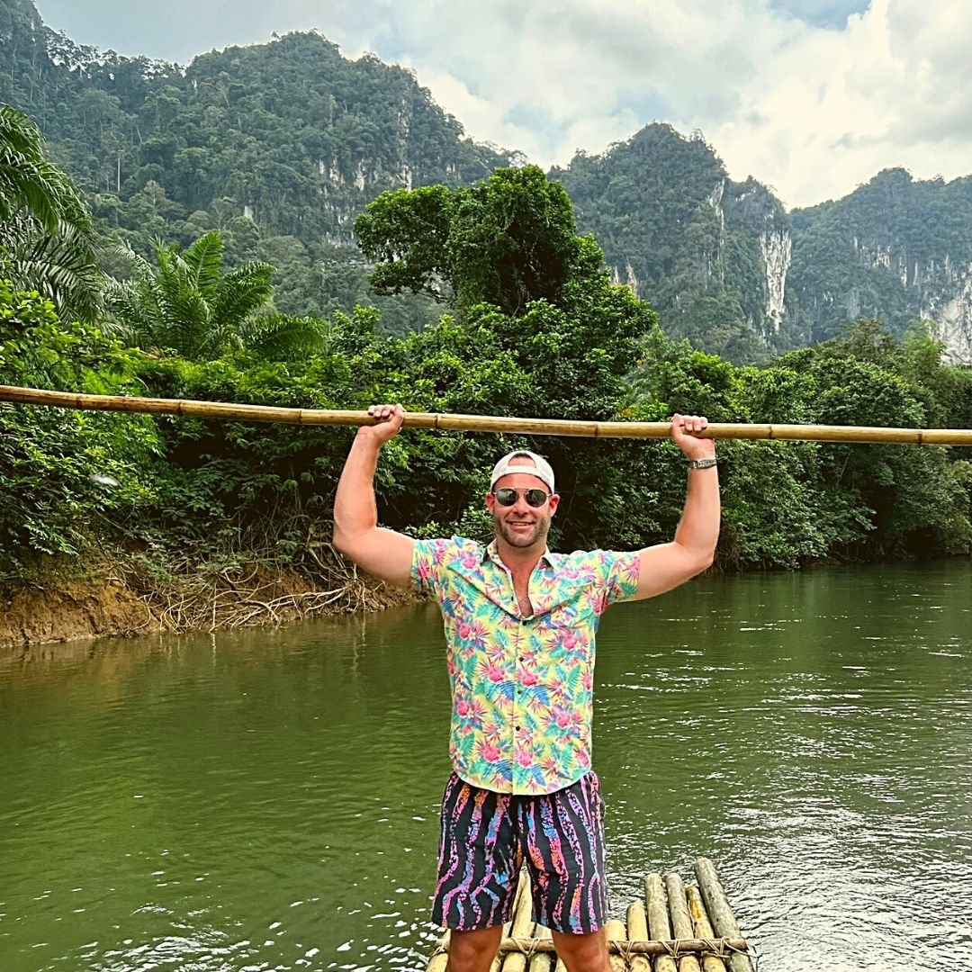 Man standing on a bamboo raft in a river with lush greenery and mountains in the background