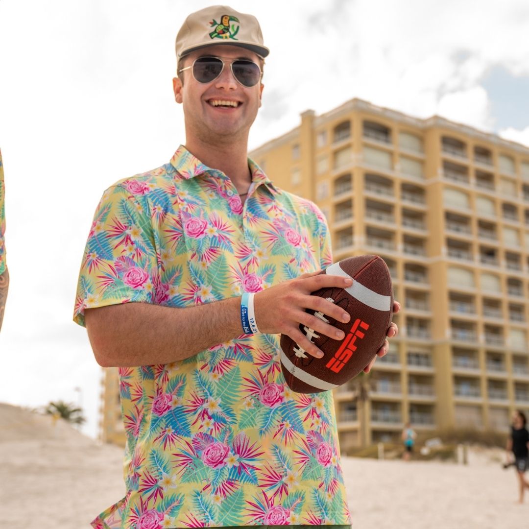 Man in a colorful shirt holding a football on a beach with a building in the background