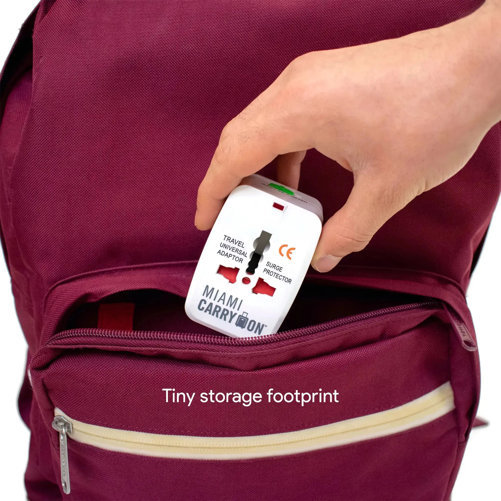 Hand placing a travel adapter into a maroon backpack with 'Miami Carry On' branding.