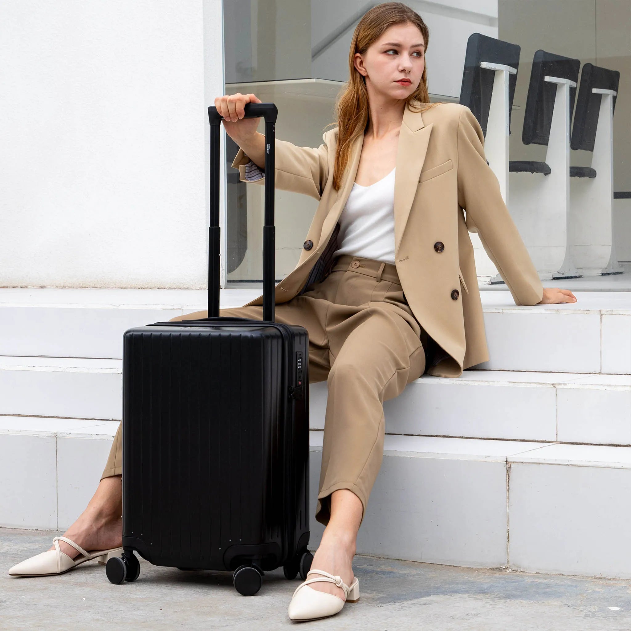 Woman in a beige suit sitting on steps with a black suitcase.