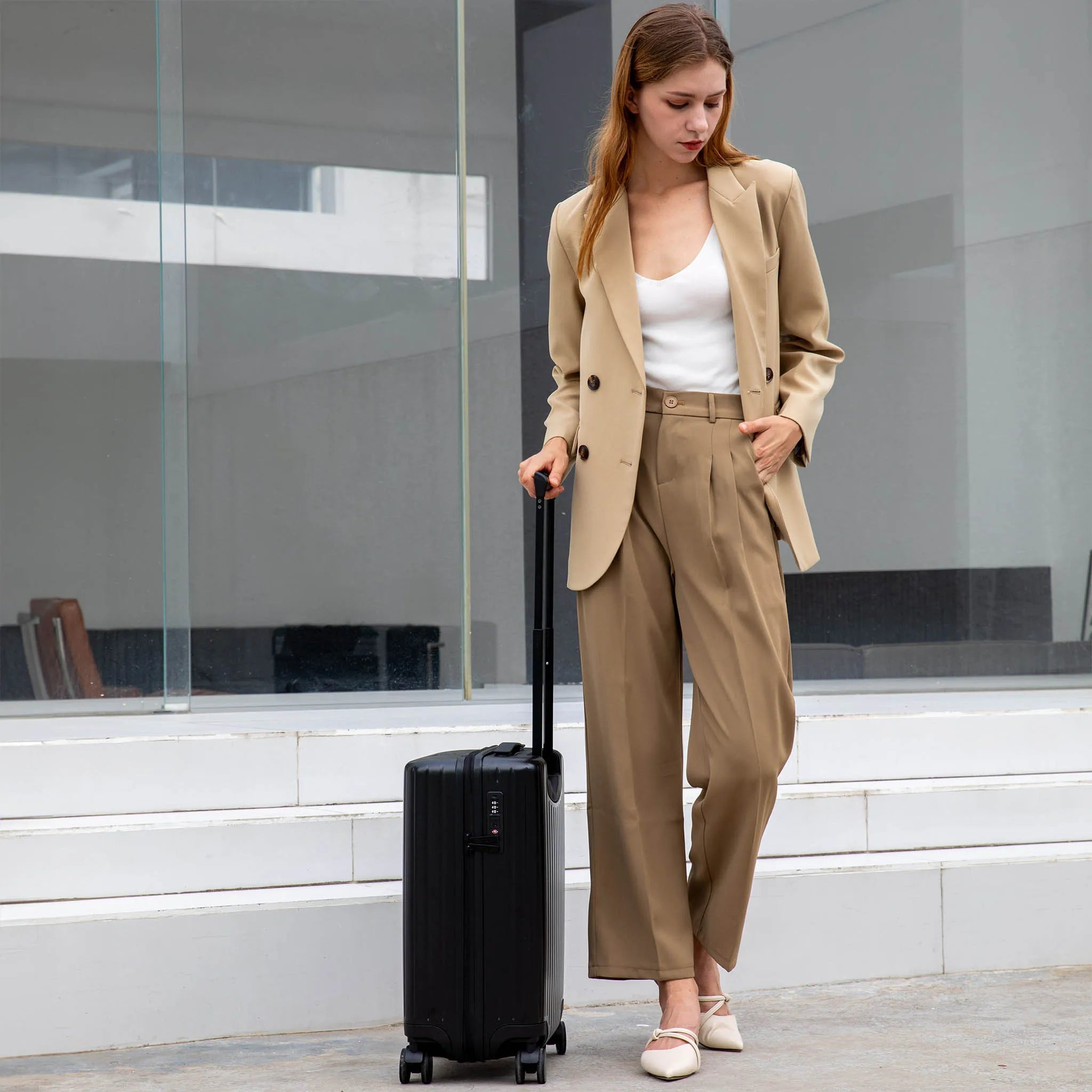 Woman in a beige suit with a black suitcase standing in front of a modern building.