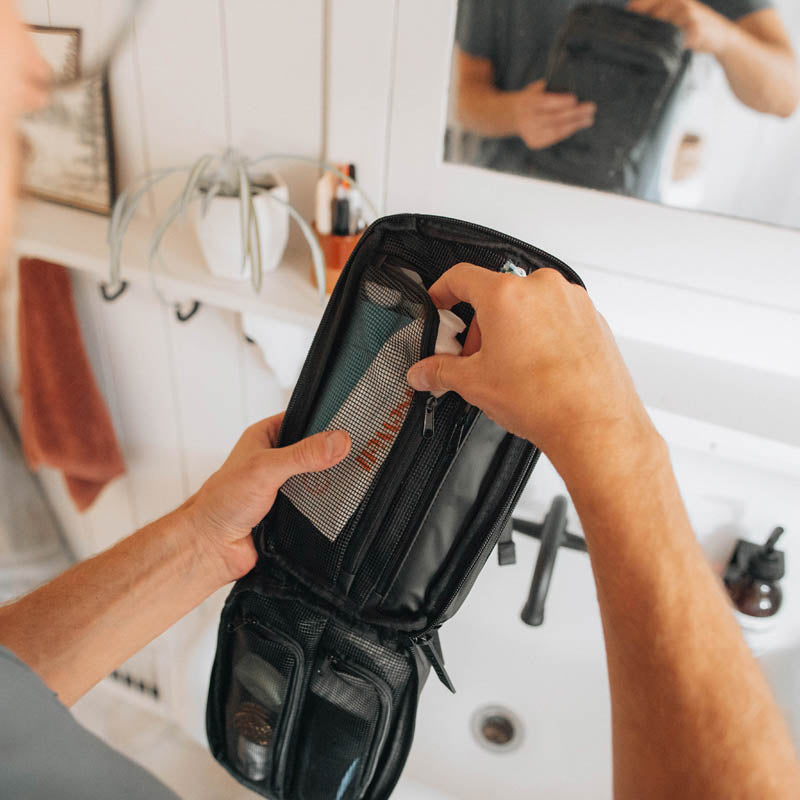 Person organizing toiletries in a black travel bag in a bathroom.