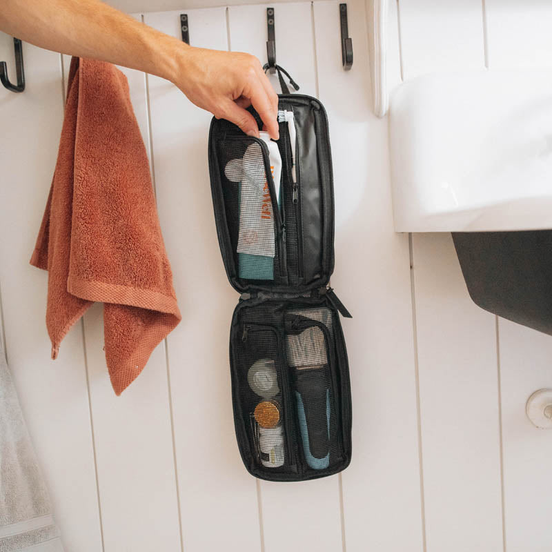 Person holding an open toiletry bag with items against a white wooden wall.