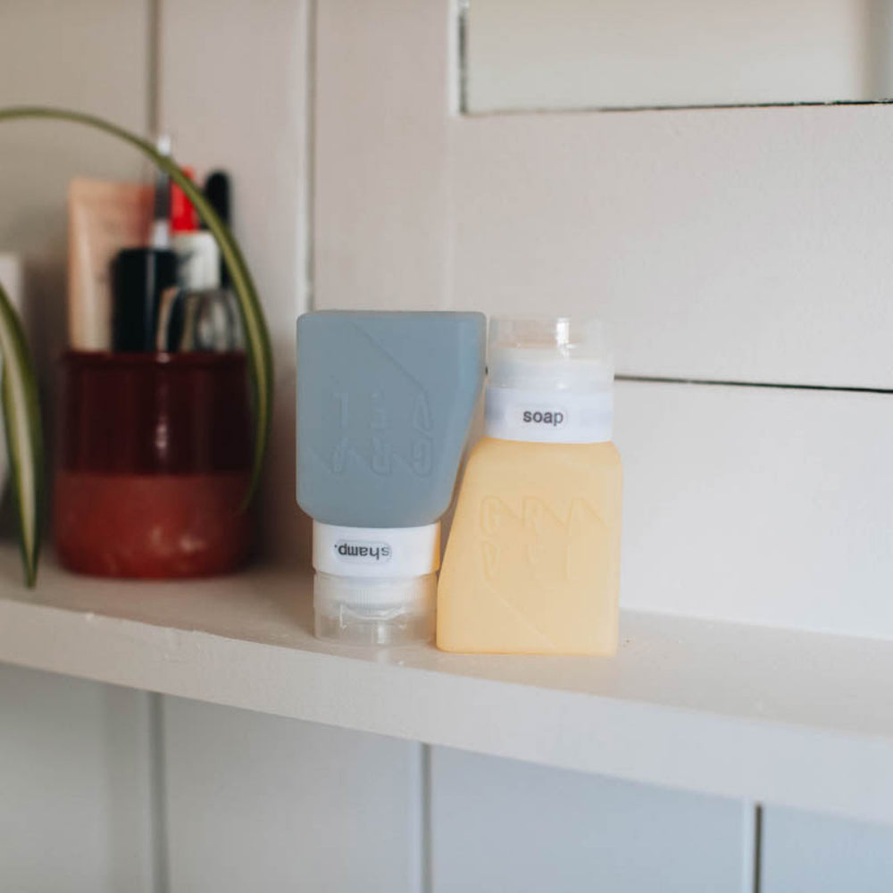 Gray and yellow soap dispensers on a shelf with a plant and other items in the background.