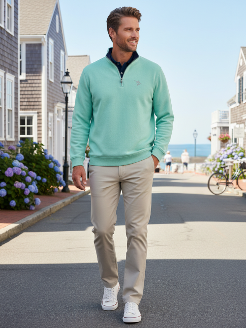 Man wearing a light green sweater and beige pants walking on a street with houses and flowers in the background.