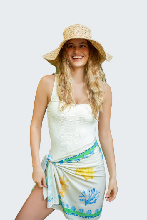 Woman wearing a white tank top and colorful sarong with a straw hat, standing in a tropical setting.