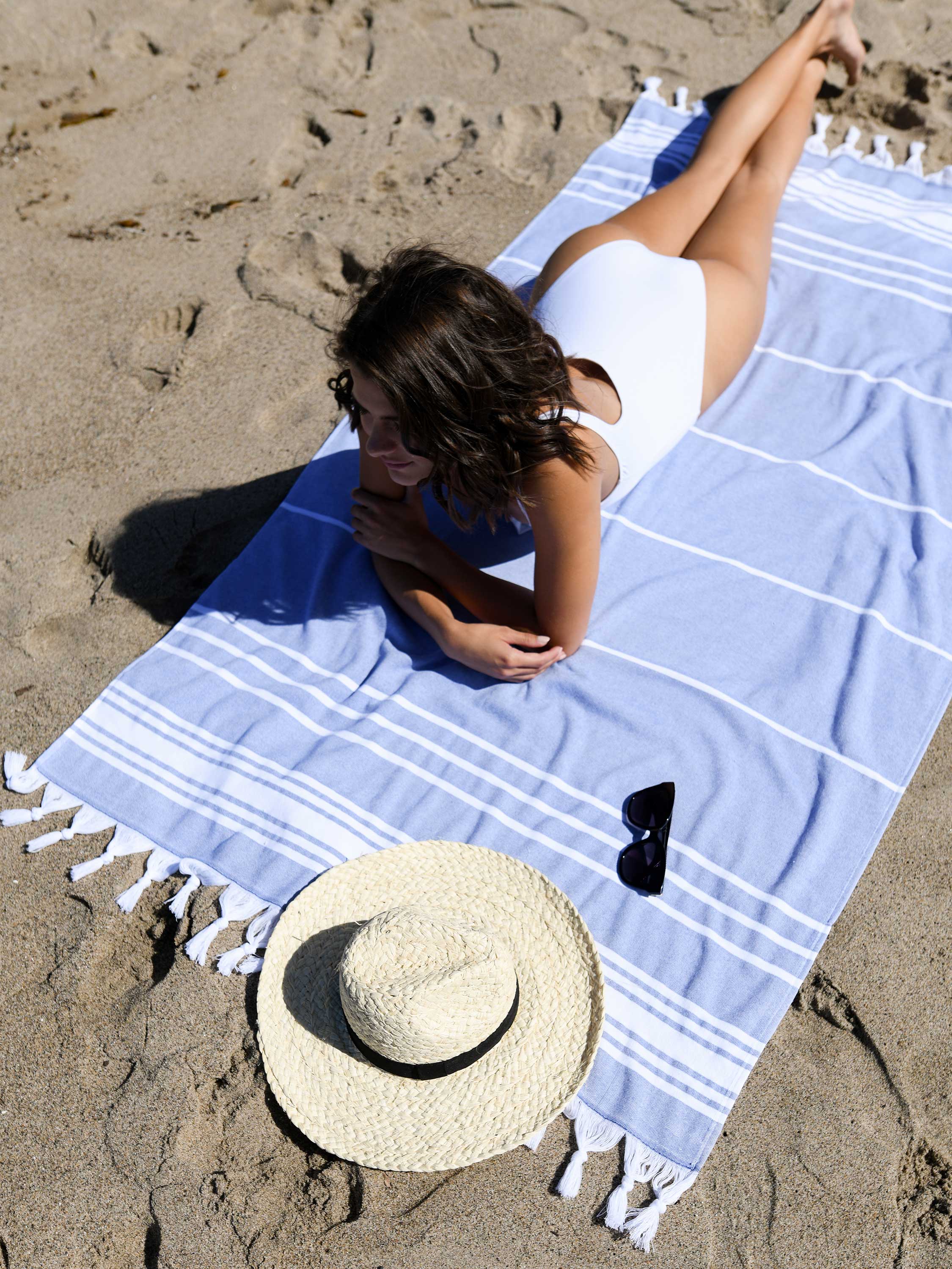 Woman lying on a blue and white striped towel at the beach with a sun hat and sunglasses nearby.
