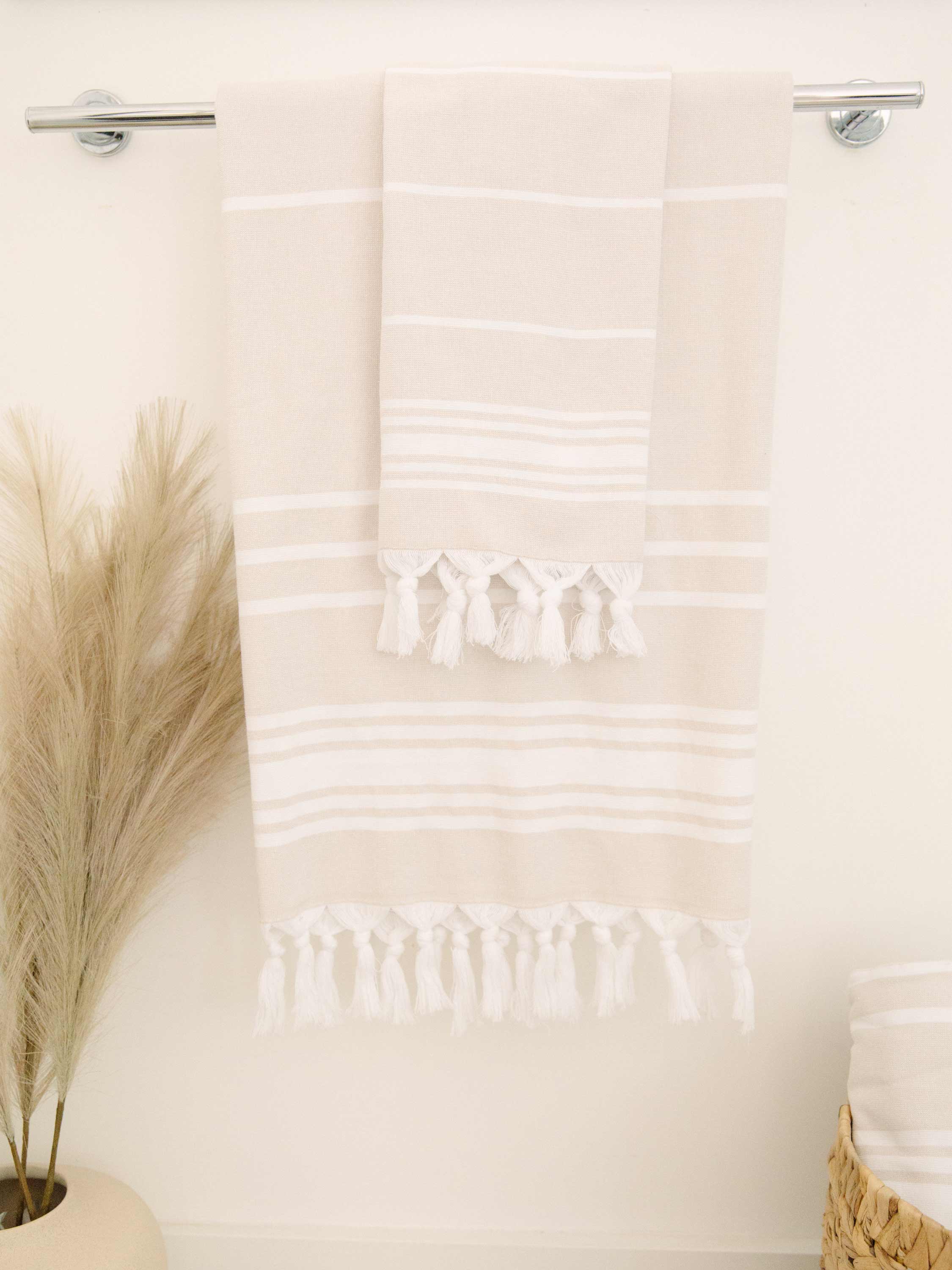 Beige towel with white stripes and tassels hanging on a rack, with decorative elements like pampas grass.