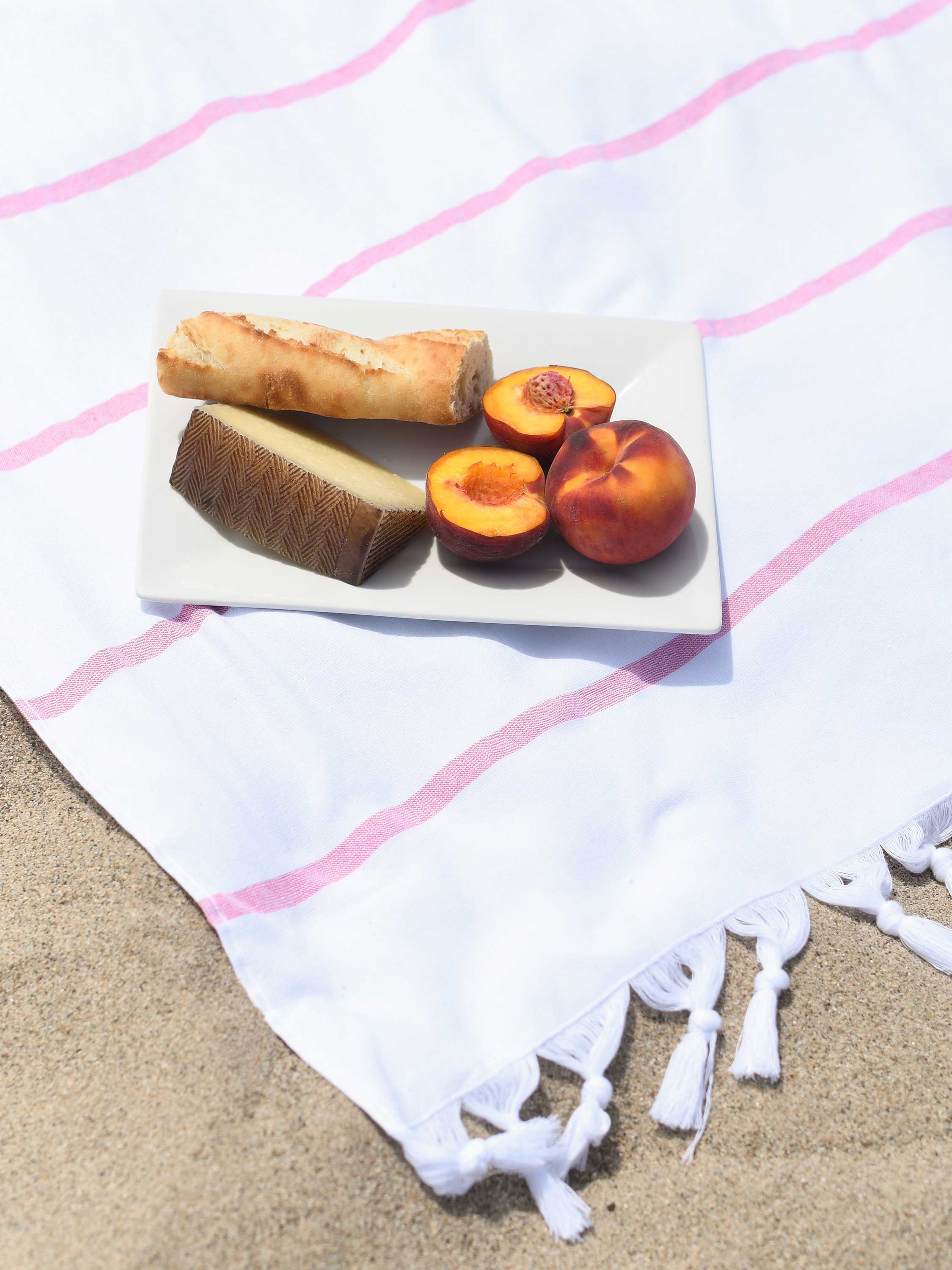 Fruit platter with peaches and a roll on a white towel with pink stripes on sand.