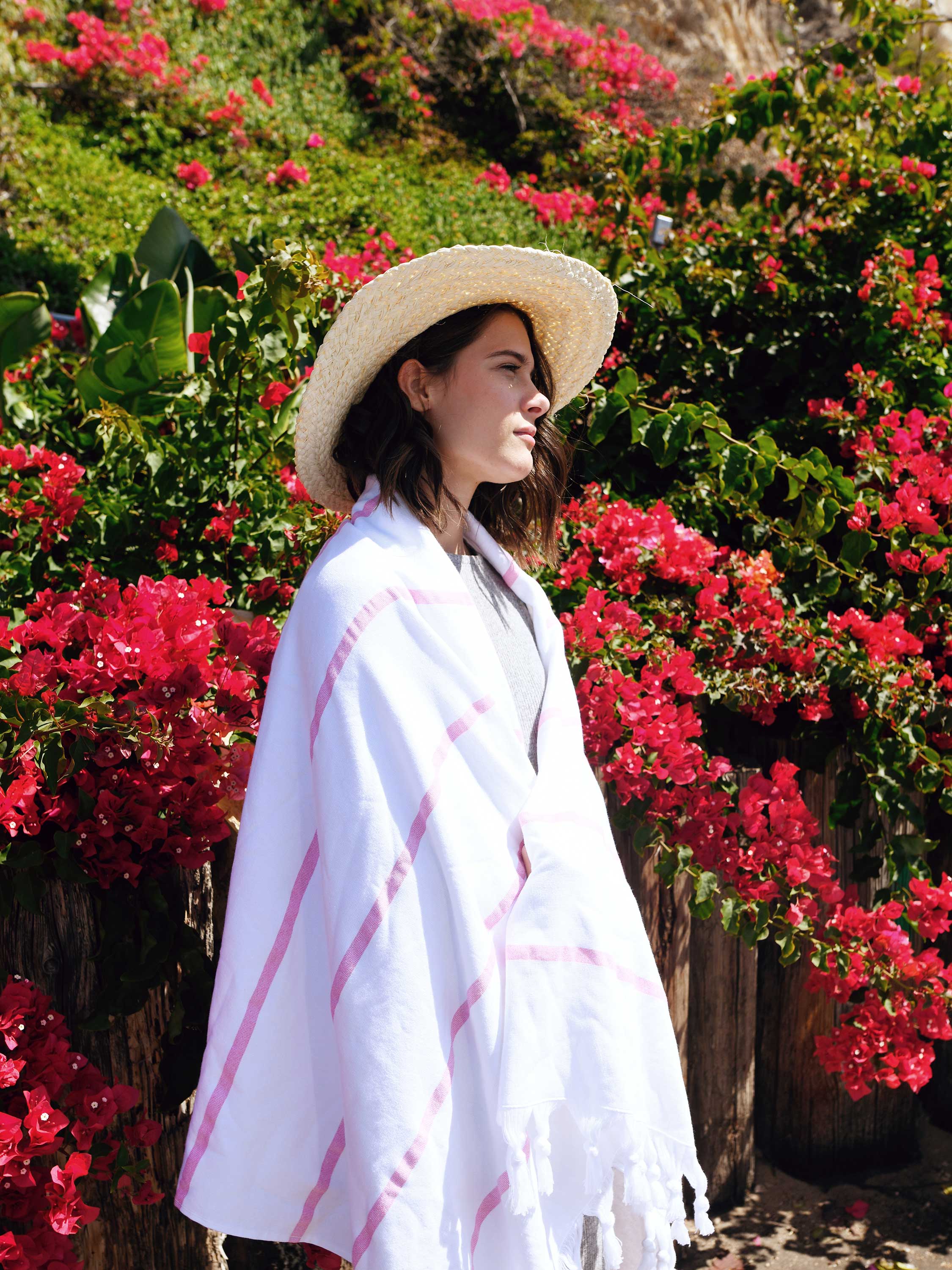 Woman wearing a white poncho with pink stripes and a straw hat, standing in front of red flowers.