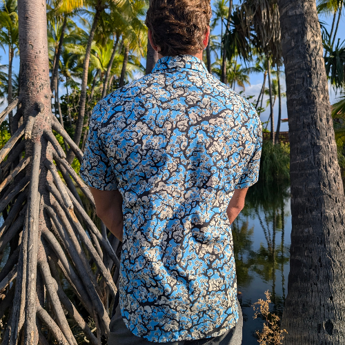 Person wearing a blue floral shirt standing among tropical trees and plants.