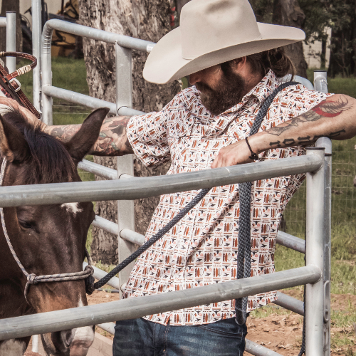 Man in cowboy hat and patterned shirt standing next to a horse in a fenced area.