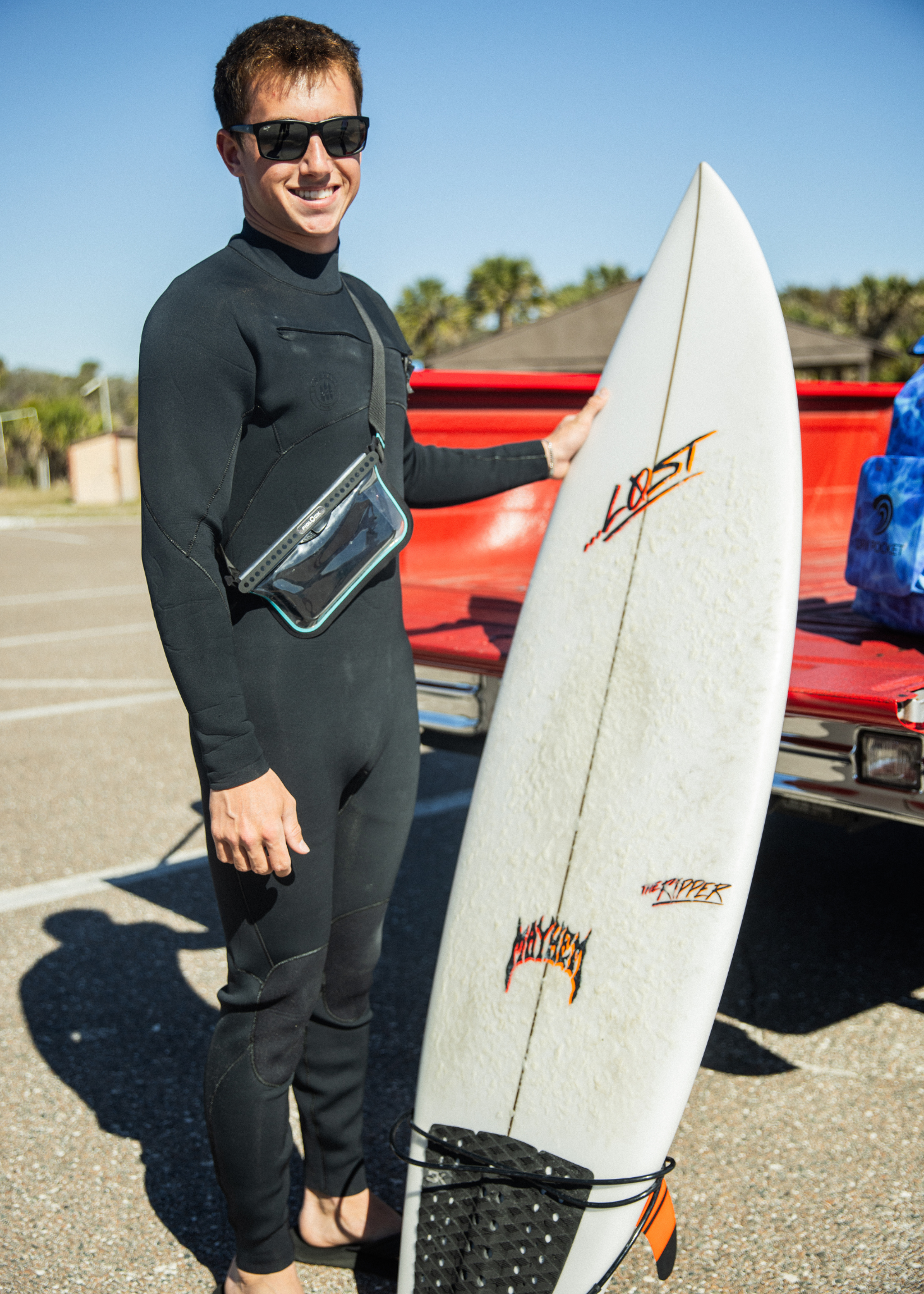 Person in a wetsuit holding a surfboard with a red vehicle in the background