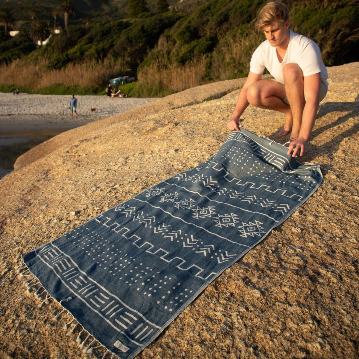 Person setting up a blue patterned towel on a rock by a beach.