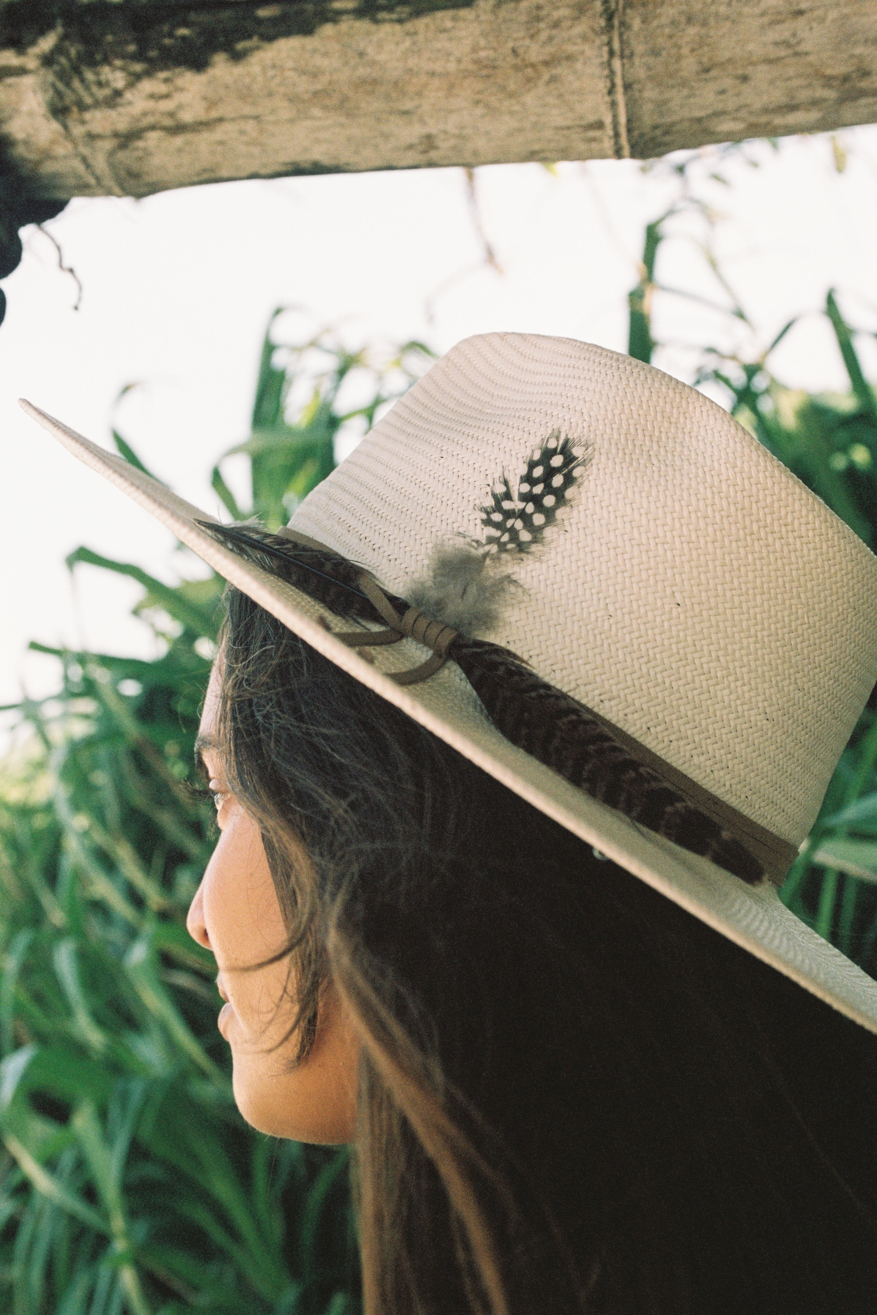 Person wearing a straw hat with a feather and pinecone decoration, standing in front of green foliage.