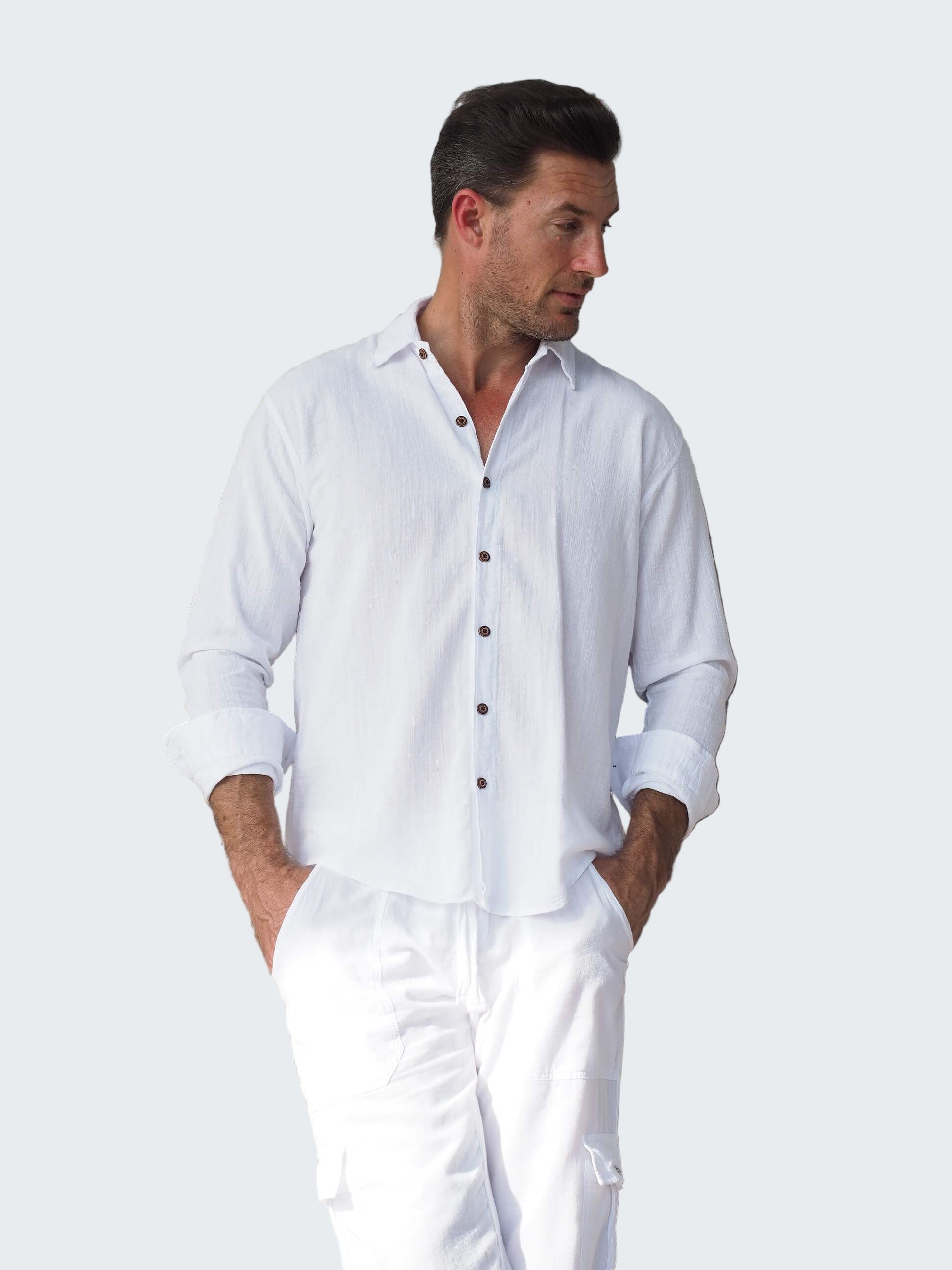 Man wearing a white shirt and pants standing against a rustic wooden wall.