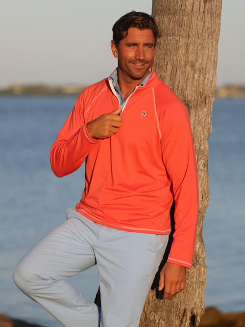 Man wearing an orange long-sleeve shirt and gray pants leaning against a tree by a body of water.