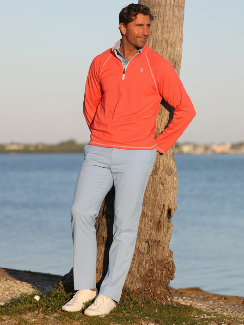 Man in an orange pullover and light blue pants leaning against a tree by a body of water.