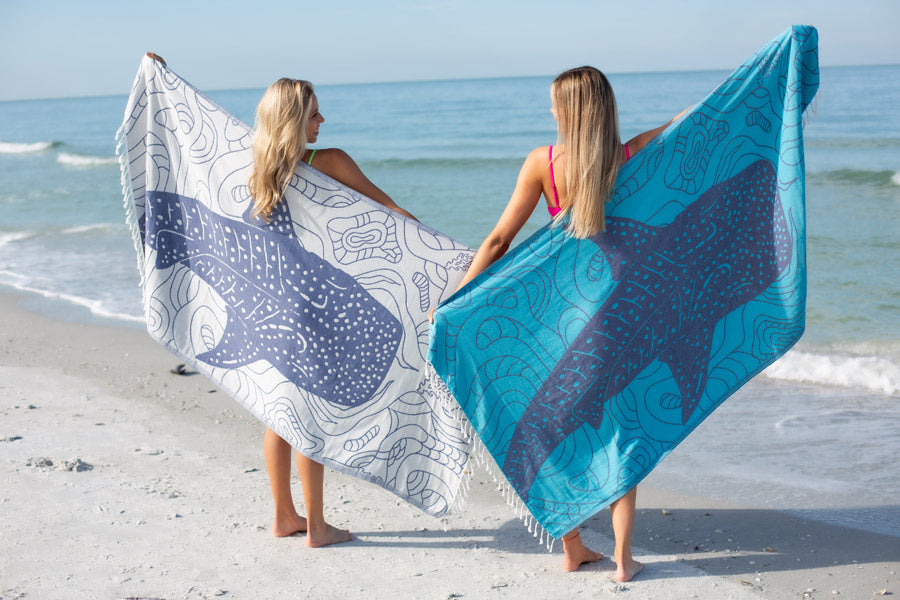 Two women on a beach holding patterned towels.